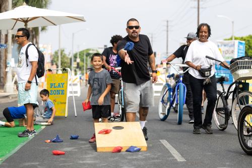 Man playing cornhole with others on a street, surrounded by bikes and people.
