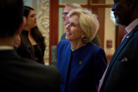 Elegant woman in blue jacket smiling, surrounded by people at an indoor event.